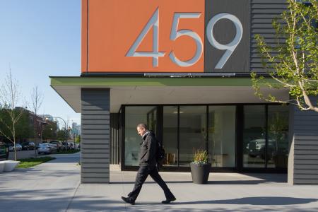 A man in a black jacket and dark pants walks on the sidewalk in front of a modern building with large glass windows. The building has a prominent orange and gray facade with the large numbers "459" displayed on it. The street is lined with trees and parked cars, and the sky is clear and blue.