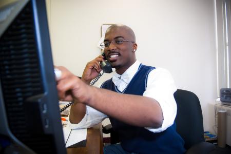 A smiling man sits at desk talking on the phone while pointing at a computer monitor.