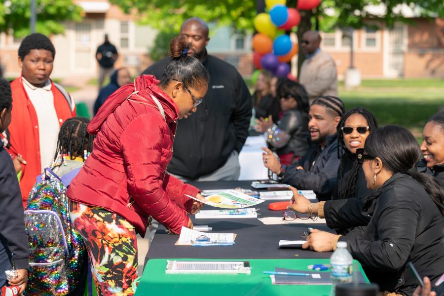 People engaging with representatives at a resource fair booth. A woman in a red jacket and colorful pants is leaning over the table, looking at a pamphlet, while others, including children and adults, are gathered around. The representatives are seated, smiling, and talking with attendees. Colorful balloons are visible in the background, indicating a lively outdoor event.