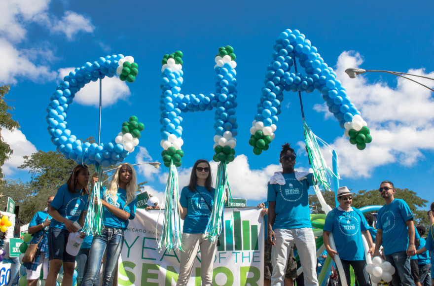 Group of people at an outdoor event holding large balloon letters that spell 'CHA.' The balloons are blue and white with green accents. The individuals, wearing blue t-shirts, stand under a bright blue sky with scattered clouds, and trees in the background. A banner behind them partially reads 'Year of the Senior.'